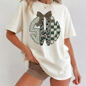 Op de afbeelding: Een crèmekleurig t-shirt met korte mouwen en een grote cirkelvormige monogramafbeelding op de borst. Het monogram bestaat uit de letters SWM, elk met een ander patroon: luipaardprint, koeienprint en een donkergroen met wit dambordpatroon.