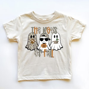 May include: A cream-colored short-sleeve t-shirt featuring a graphic of three ghosts. The text reads "THE BOYS OF FALL". One ghost has a camouflage pattern and holds a football, another wears sunglasses and a cap while holding a pumpkin, and the third holds a mallard duck.