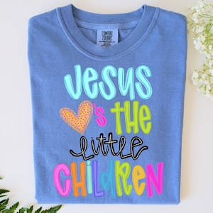 May include: A blue Comfort Colors t-shirt with the words "Jesus loves the little children" in colorful, playful lettering. A yellow heart with pink polka dots is also featured on the shirt.