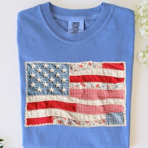 May include: A light blue t-shirt with a sewn-on American flag design. The flag features white stars on a blue denim field and red and white striped sections made of various patterned fabrics. The shirt has a crew neck and the brand name "Comfort Colors" is visible.