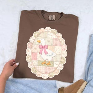 May include: A brown t-shirt featuring a white goose with a pink bow, set within an oval frame with a patchwork design. The goose has orange feet and beak. The shirt is a solid brown color.