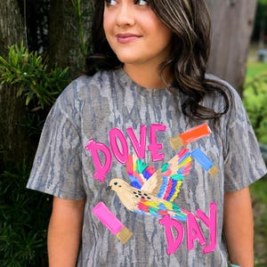 Puede incluir: Una camiseta con estampado de camuflaje gris que presenta un gráfico de una paloma con alas multicolores. El texto "DOVE DAY" está impreso en letras de color rosa brillante. Tres cartuchos de escopeta en naranja, azul y rosa rodean al ave.