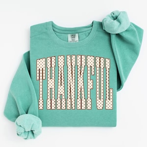 May include: A teal crewneck sweatshirt featuring the word THANKFUL printed in a large, arched, cream-colored font with brown polka dots and a brown outline. The garment has a Comfort Colors brand label visible at the inner neckline.