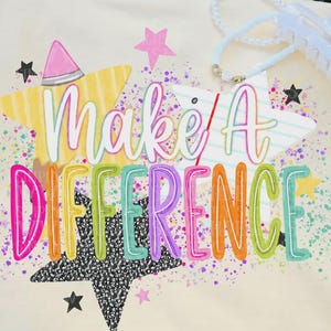 Peut inclure: Un t-shirt couleur crème arborant un motif coloré avec le texte "Make A DIFFERENCE" dans une police multicolore. Le design comprend des étoiles jaunes et blanches, une pointe de crayon rose et des étoiles noires mouchetées avec des éclaboussures violettes.