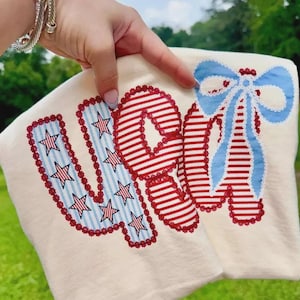 Könnte beinhalten: Off-white Stoff mit dem Wort "VOTE" in rot, weiß und blau appliziert. Das "V" hat blau-weiß gestreiften Stoff mit roten Sternen. Das "O" und "E" sind rot-weiß gestreift. Eine blaue Schleife befindet sich auf der rechten Seite.
