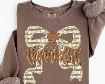 May include: A brown long-sleeve sweatshirt featuring a graphic of a large beige and white gingham bow with a brown leaf at the center. The word "thankful" is printed in a cursive, orange-brown script across the center of the bow design.