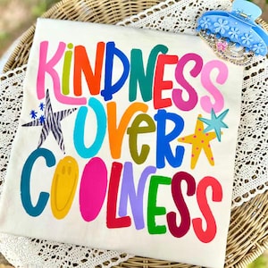 May include: A cream-colored t-shirt with the words "KINDNESS OVER COOLNESS" in vibrant, multicolored letters. The design includes star and smiley face graphics. A blue hair clip with flower details is in the upper right corner.