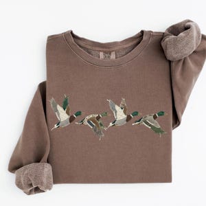 May include: A brown crewneck sweatshirt featuring a graphic of four flying mallard ducks. The ducks are depicted with a patchwork-style design in various earth tones and patterns. The inner neck label displays the text Comfort Colors.