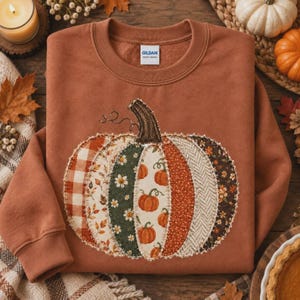 Op de afbeelding: Een roestkleurige sweater met ronde hals en een grote, centrale pompoenafbeelding die is samengesteld uit verschillende stofachtige vlakken. Het ontwerp bevat segmenten met ruitjes, bloemen en bladeren in tinten oranje, groen en crème.