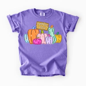 May include: A purple short-sleeve t-shirt featuring a graphic of various colorful pumpkins in shades of orange, pink, yellow, blue, and green. A wooden-style sign above the pumpkins displays the text PUMPKIN PATCH in white lettering.