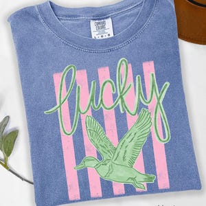 May include: Blue jean coloured t-shirt with a graphic design. The design features the word "lucky" in green script over pink vertical stripes, and a green duck in flight. The shirt has a crew neck and short sleeves.