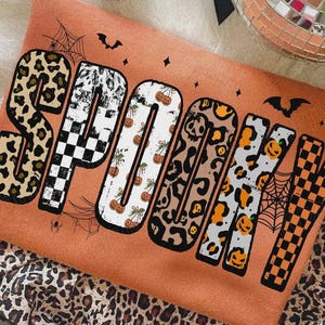 Può includere: Una felpa arancione con la parola SPOOKY in grandi lettere stampatello. Ogni lettera presenta un motivo diverso, tra cui stampa leopardata, scacchiera bianca e nera, zucche e ragnatele. Piccoli pipistrelli e stelle sono stampati attorno al testo.