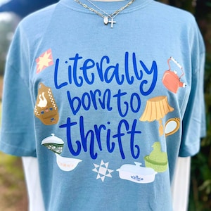 Op de afbeelding: Lichtblauw T-shirt met de tekst "Literally born to thrift" in blauw schrift. Het shirt heeft kleurrijke illustraties van verschillende tweedehands items, waaronder een theepot, lamp en ovenschaal. Een ketting met een kruisvormige hanger is zichtbaar.