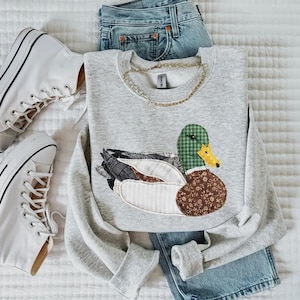 May include: A light gray sweatshirt featuring a duck applique with green plaid, floral, and patterned fabric details. The sweatshirt is paired with blue jeans and white high-top sneakers, and a gold chain necklace.