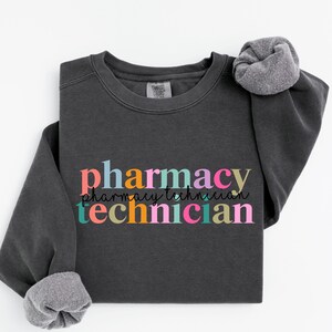 May include: A charcoal gray crewneck sweatshirt featuring the word pharmacy in colorful block letters above the word technician in matching colors, with the words pharmacy technician in a thin black cursive script layered in between.