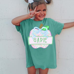 May include: A teal short-sleeve t-shirt featuring a graphic of an apple with a light blue bow. The apple has a speckled pattern in pink, green, and white. A rectangular banner across the center of the apple contains the name SADIE in green letters.