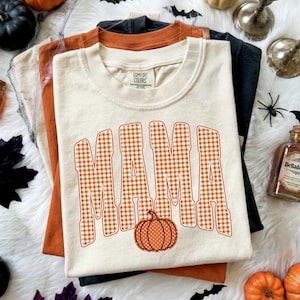 May include: A cream-colored short-sleeve t-shirt featuring the word MAMA in a large orange and white gingham pattern. Below the text is a small orange pumpkin with white polka dots. The shirt has a Comfort Colors brand label at the neck.