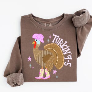 May include: A brown crewneck sweatshirt featuring a graphic of a turkey wearing a pink cowboy hat and pink cowboy boots. The text "Turkin' 9-5" is printed in light pink next to the turkey, accompanied by small pink and blue star icons.