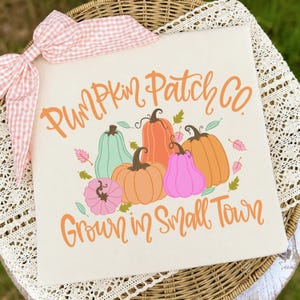 May include: A square white sign featuring a pink gingham bow at the top left corner. The text reads PUMPKIN PATCH CO. and GROWN IN SMALL TOWN in orange script. The center displays a cluster of illustrated pumpkins in orange, pink, and mint green.