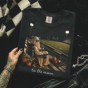 May include: A black t-shirt featuring a graphic of a ghost wearing a sheet and sitting in a folding chair on a football field at night. The ghost holds a coffee cup, surrounded by pumpkins and string lights. The text 'tis the season' is printed below.
