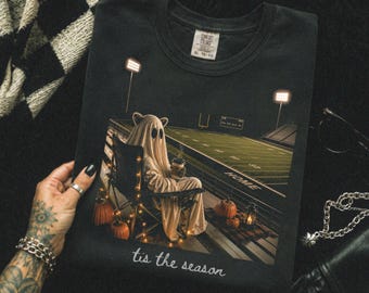 May include: A black t-shirt featuring a graphic of a ghost wearing a sheet and sitting in a folding chair on a football field at night. The ghost holds a coffee cup, surrounded by pumpkins and string lights. The text 'tis the season' is printed below.