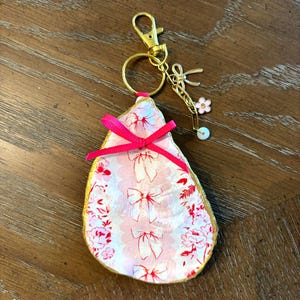 May include: A decorative seashell keyring with a pink and white floral pattern and a gold-coloured trim. The shell is adorned with a bright pink bow and a gold-coloured clasp, key ring, and charms, including a flower and a pearl.
