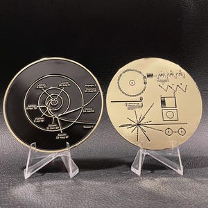 May include: Two round Voyager Golden Record replicas on clear stands. One is black with a diagram and text, the other gold with a diagram. The black disc shows a spiral and dates. The gold disc has lines and symbols.