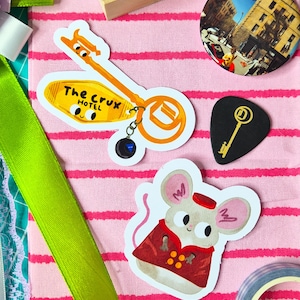 Può includere: Una collezione di adesivi e un bottone su sfondo rosa a righe. Gli adesivi includono una chiave d'albergo con il testo "The Crux Hotel", un topo con cappotto rosso e un plettro nero con design a chiave dorata. Un bottone raffigura una scena cittadina.