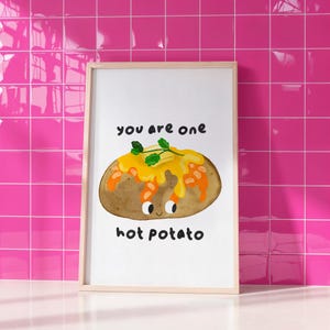 Może przedstawiać: Oprawiony wydruk z akwarelową ilustracją pieczonego ziemniaka z serem i pietruszką. Tekst "you are one hot potato" jest napisany nad i pod ziemniakiem. Rama jest z jasnego drewna.