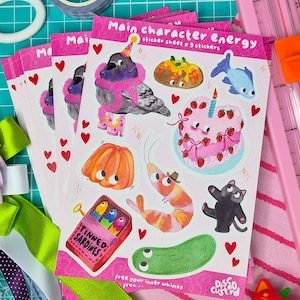 May include: Sheets of stickers with the text "Main character energy" featuring whimsical illustrations of a pigeon, cake, and other characters. The stickers are pink with red heart accents and the text "Tinned Sardines".