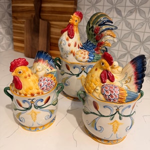 May include: Three ceramic canisters with lids featuring sculpted roosters and hens. Each cream-colored jar has two green handles and hand-painted floral scrollwork in blue, green, and yellow. The lids are topped with detailed poultry figures.