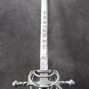 Peut inclure: Une épée de rapière argentée avec un manche noir. La lame présente des marquages gravés et une pointe acérée. La garde présente un design complexe. L'épée est présentée sur un fond sombre.