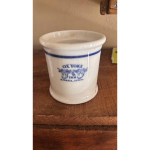 May include: A white ceramic mug with a blue band and text. The text reads "OX YOKE INN AMANA, IOWA" with a graphic of two oxen. The mug has a slightly flared rim and a rounded base.
