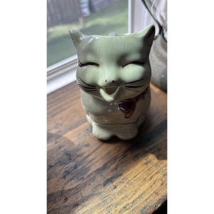 May include: A ceramic cat figurine with a smiling face, closed eyes, and black whiskers. The cat is light green with a dark red bow around its neck. The figurine is sitting on a wooden surface.