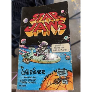 Puede incluir: Un cómic vintage titulado "Star Jaws" con un fondo negro e ilustraciones coloridas. La portada presenta un robot en una nave espacial, planetas y el texto "Stupid... That's the Wrong Planet!" El autor es Will Eisner.