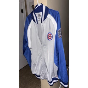 Może przedstawiać: Niebieska, biała i szara kurtka baseballowa Chicago Cubs. Kurtka ma pełny zamek błyskawiczny, logo Cubs na piersi i naszywkę na rękawie. Kurtka wisi na białym wieszaku.