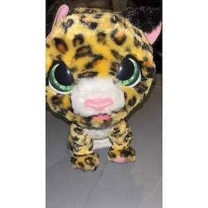 Op de afbeelding: Een pluche luipaard met een geel en zwart gevlekt patroon. Het heeft grote groene ogen, een roze neus en een roze tong. De oren zijn zwart met roze binnenkant. Het speelgoed is ontworpen om op een baby luipaard te lijken.
