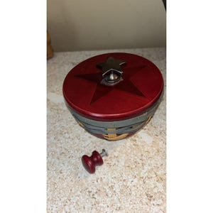 Pode incluir: Uma cesta decorativa redonda vermelha com um desenho de estrela na tampa. A tampa tem uma alça prateada em forma de estrela. A cesta tem faixas tecidas cinza e amarelas nas laterais. Um pequeno botão vermelho está ao lado da cesta.