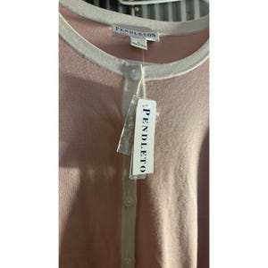 May include: A light pink cardigan with a cream-colored trim and buttons. The tag reads "PENDLETON" at the top and on the attached label. The cardigan is made of a soft material and has a classic design.