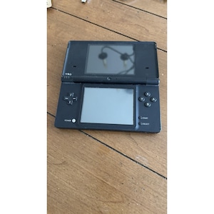 May include: Black Nintendo DS handheld game console, open and displayed on a wooden surface. The console features two screens, a directional pad, and action buttons. The top screen reflects the surroundings.