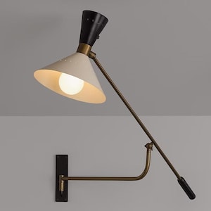 Könnte beinhalten: Eine Wandleuchte im Mid-Century-Modern-Stil mit weißem, konischem Schirm und schwarzem Oberteil. Die Lampe hat einen messingfarbenen Arm und eine schwarze Montageplatte. Das Licht ist an und beleuchtet das Innere des Schirms.