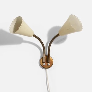 May include: A vintage wall-mounted lamp with two cream-colored, cone-shaped shades. The shades have a scalloped edge and are attached to flexible, dark brown metal arms. The lamp has a brass-colored base with two small switches and a white power cord.