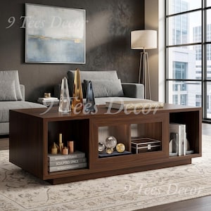 May include: A rectangular dark wood coffee table featuring open storage compartments and a central glass-fronted cabinet. The table holds stacked books, decorative glass obelisks, spherical ornaments, and a small box, with the text 9 Tees Decor visible.