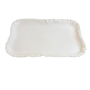 May include: A rectangular white ceramic serving tray featuring a smooth, flat center surface and a decorative, scalloped edge with subtle embossed detailing along the entire perimeter. The tray has a glossy finish and rounded corners.