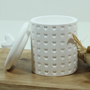 Può includere: Una tazza in ceramica bianca con manico a forma di cuore e intagli a forma di cuore. Un piccolo bouquet di fiori secchi e un fiore bianco sono accanto alla tazza su una superficie di legno. La tazza è cilindrica.