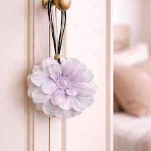 Op de afbeelding: Een decoratief bloemenornament in wit- en lavendeltinten, hangend aan een goudkleurige knop op een witte kastdeur. De bloem is gemaakt van gelaagde bloemblaadjes en wordt opgehangen door zwarte, witte en beige koorden.