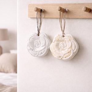 Puede incluir: Dos bombas de baño blancas en forma de rosa colgadas de un perchero de madera. Una es blanca, la otra tiene motas doradas. Las bombas de baño están suspendidas por hilo. El fondo muestra un dormitorio con una cama y una lámpara.