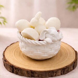 May include: A white ceramic basket filled with white bunny and egg-shaped decorations. The basket sits on a round, wooden slice, creating a rustic and festive display. Perfect for Easter or spring decor.