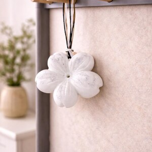 May include: A white, flower-shaped decorative item with silver accents hangs from a wooden rack. The rack is mounted on a light-colored wall. The flower is suspended by black and white cords.