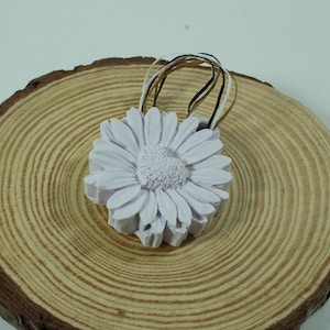 May include: A pale purple daisy-shaped ornament with a heart-shaped centre, on a wooden slice. The ornament has a loop made of black, white, and tan ribbons. The wood slice shows visible growth rings.